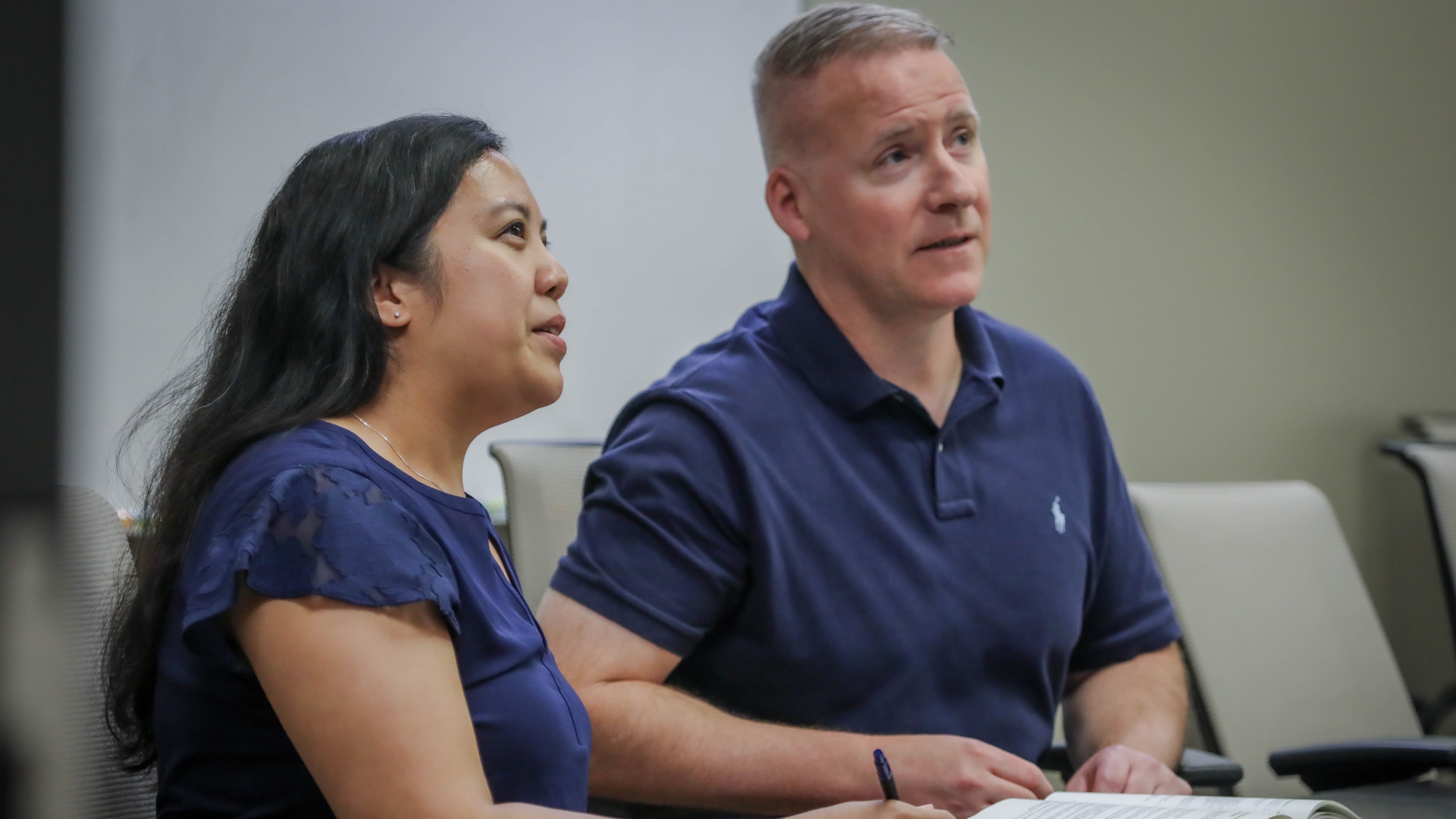 man and  woman wearing blue shirts, sitting at a table, looking up as though at someobody off camera