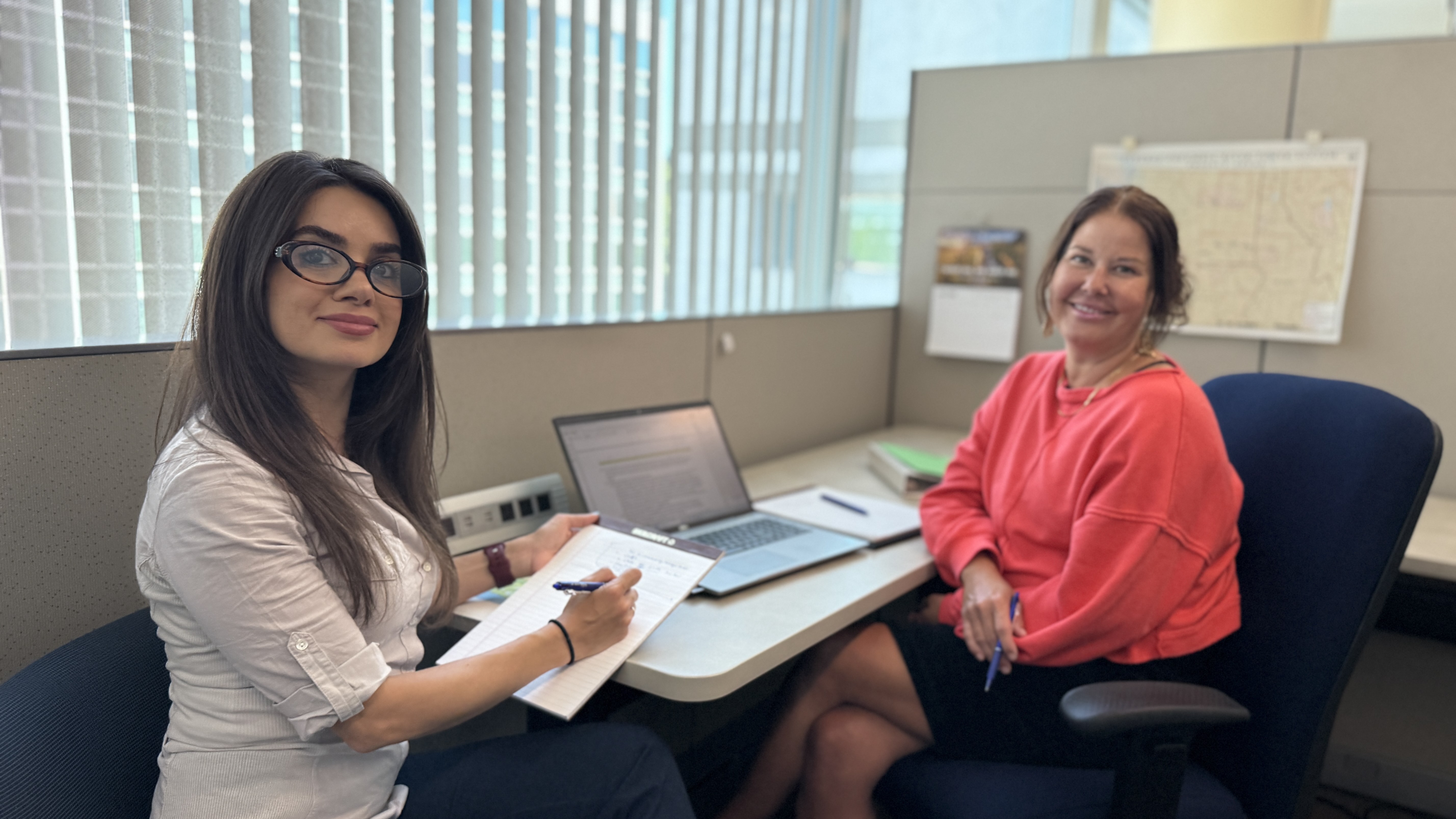two women sitting at a desk with a laptop between them, smiling 
