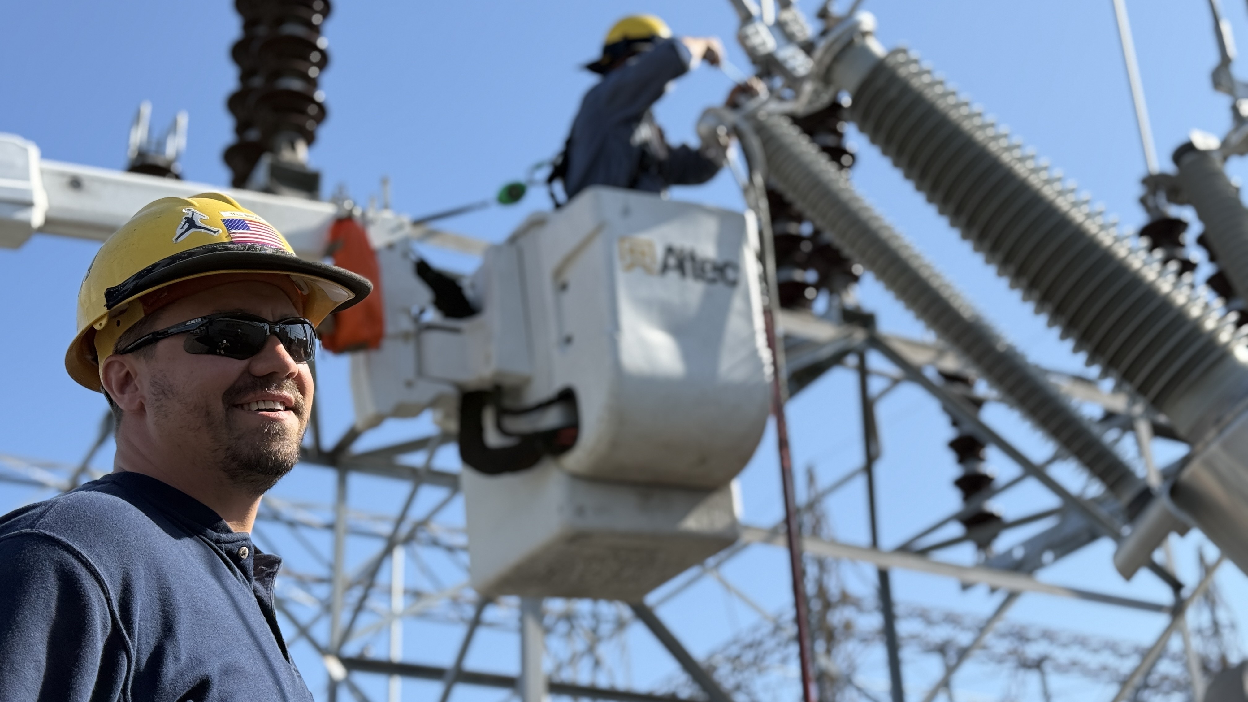 man with hardhat and sunglasses standing in front of electrical equipment and man in bucket