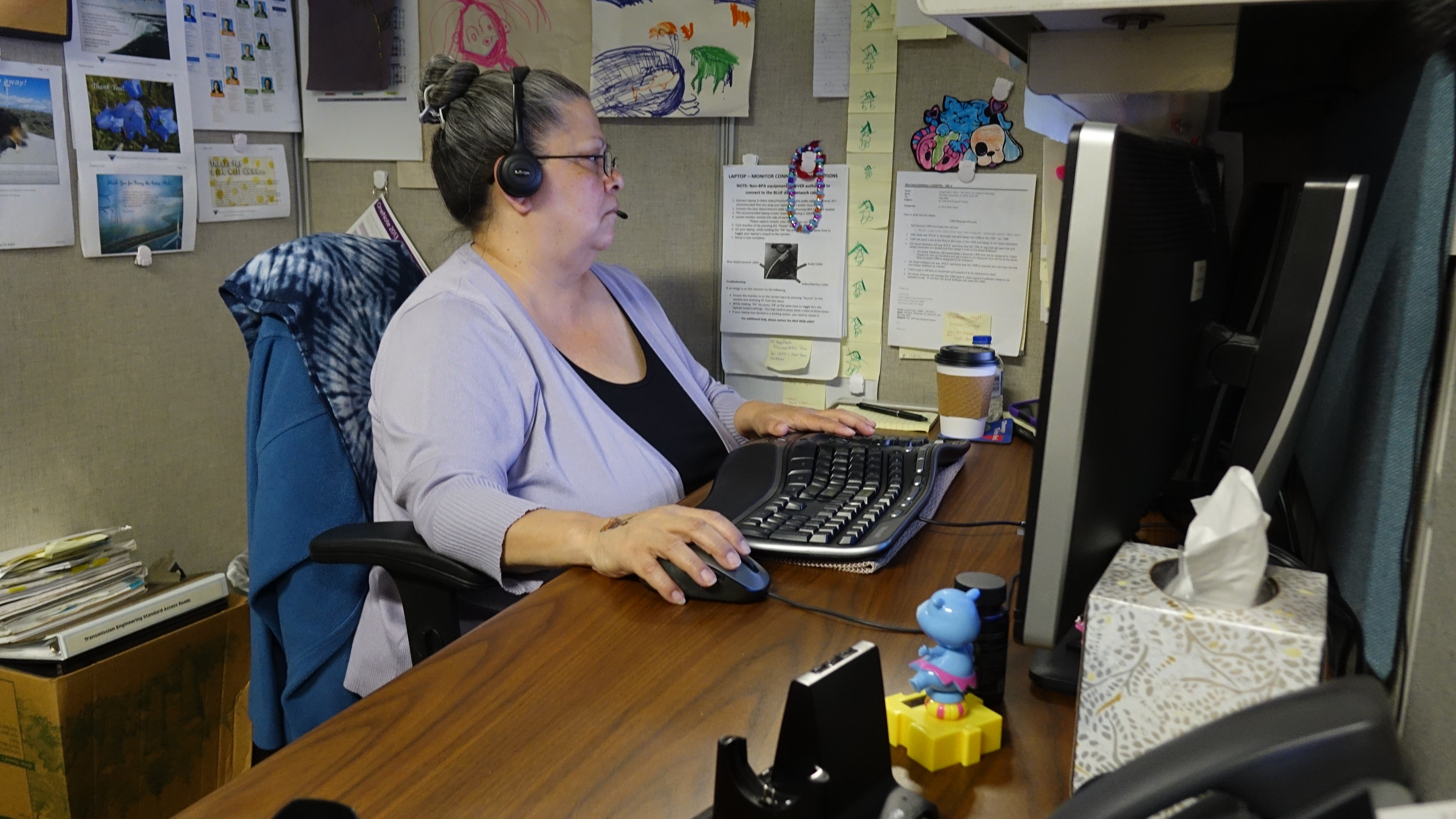 woman sitting at a desk, wearing a headset, looking at a monitor, with one hand on a mouse and the other near a keyboard