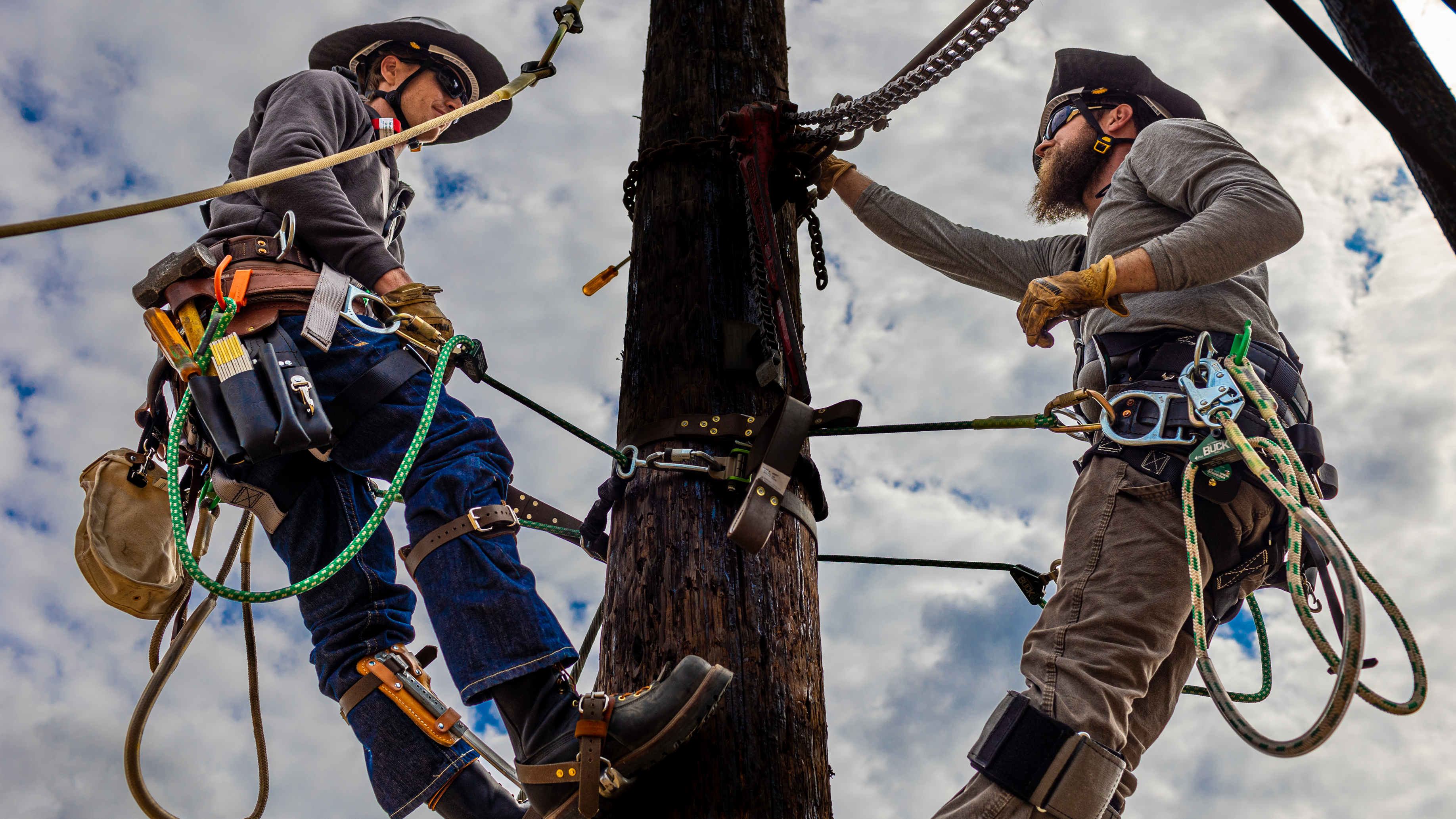 two men hanging off of electrical equipment, wearing safety gear