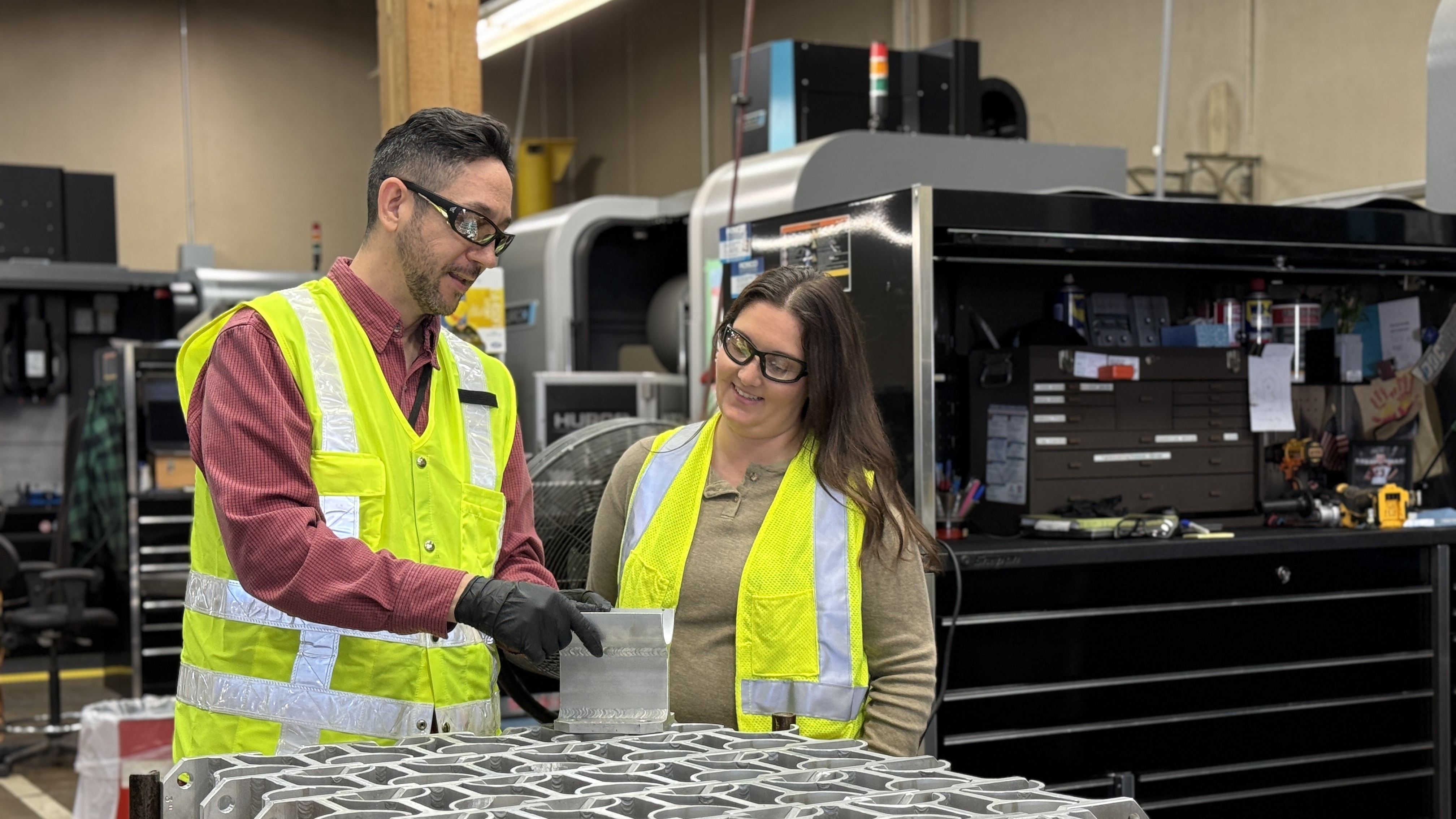 man and woman standing in a room, wearing fluorescent vests and safety glasses, standing next to a table with grey blocks on  it, with equipment in the background