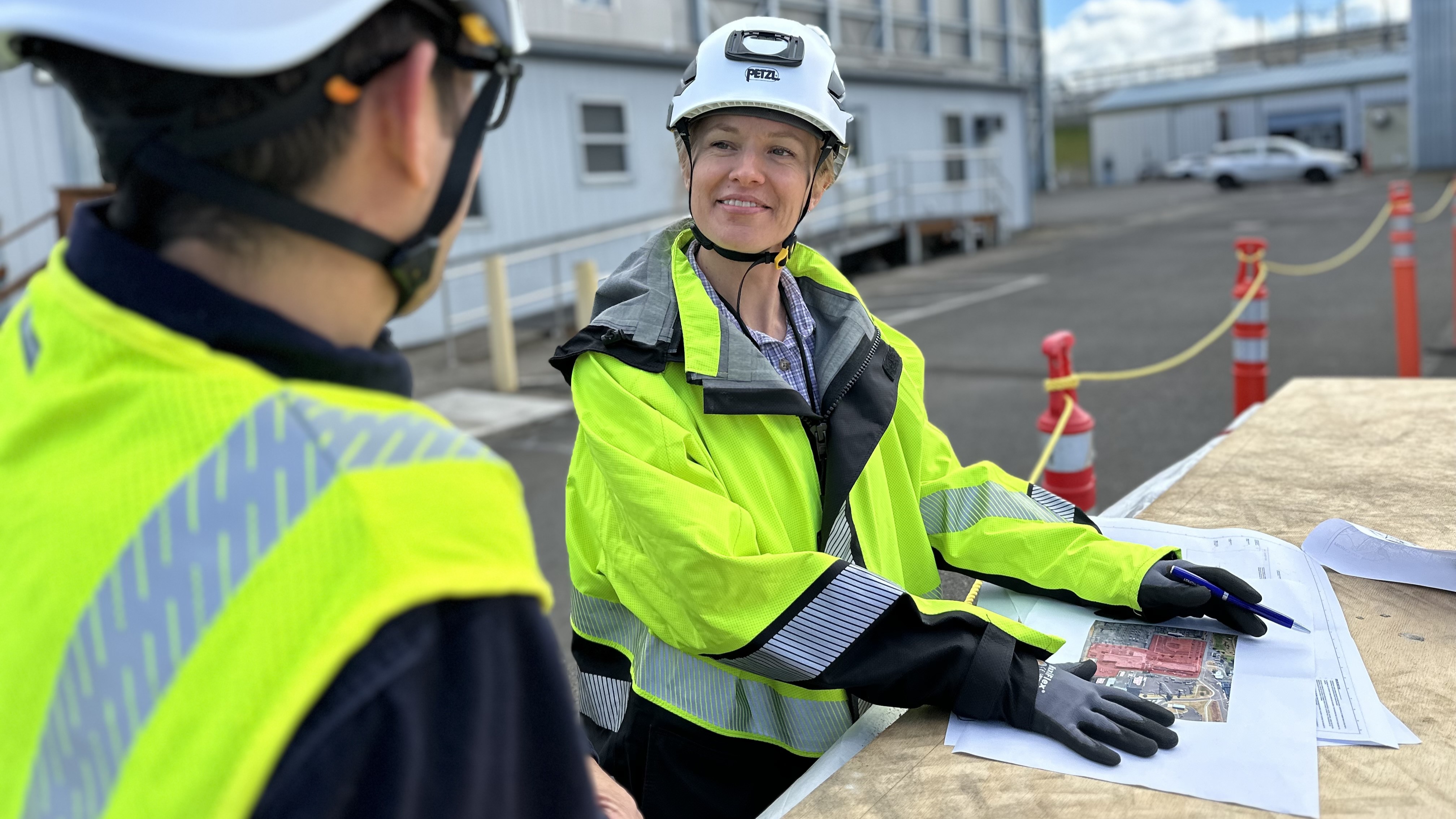 two people looking at each other, over a table with paper plans on it, the back of one person and the front of another person, both of which are wearing hard hats and fluorescent safety jackets ,with buildings and vehicles in the background