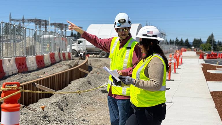 man pointing, next to woman holding paper, both wearing fluorescent yellow safety vests, standing on a sidewalk adjoining gravel and cones