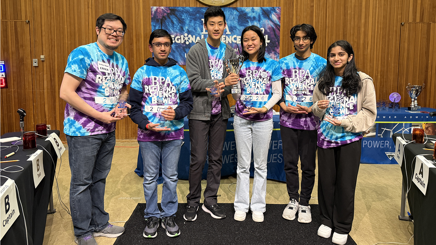 group of people wearing matching science bowl t-shirts, one of which is holding a trophy